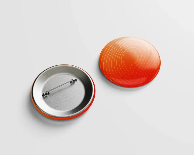 Dos escarapelas de botón: una naranja con líneas concéntricas y otra de espaldas, con el gancho metálico.