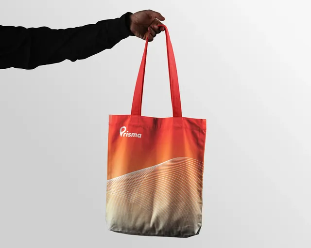 Una mano sostiene una tote bag con degradado naranja y el logotipo de Prisma.