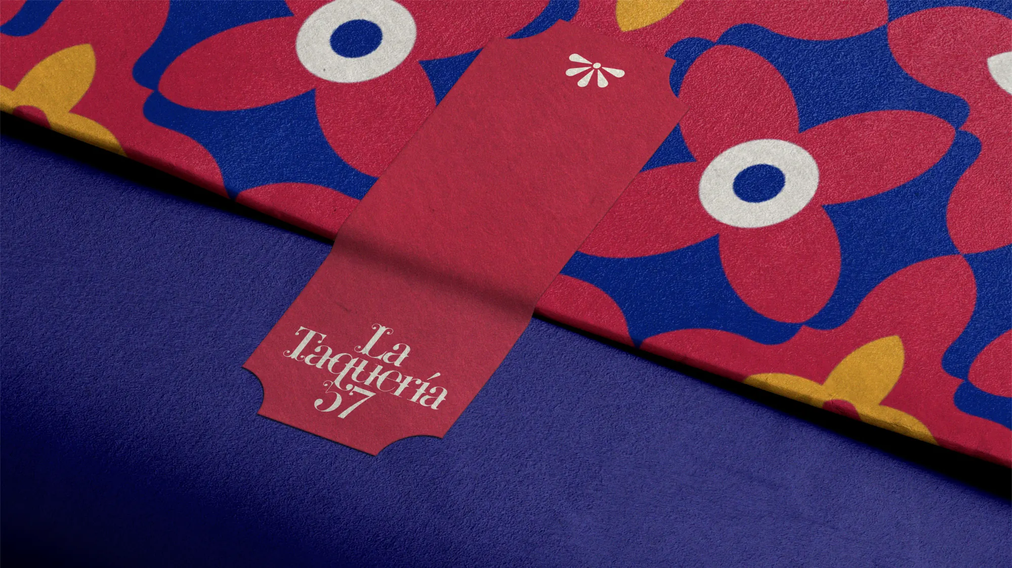 Patrón tipo talavera de La Taquería 57 en rojo, azul cobalto y mostaza, con una etiqueta colgante roja con el wordmark de la marca