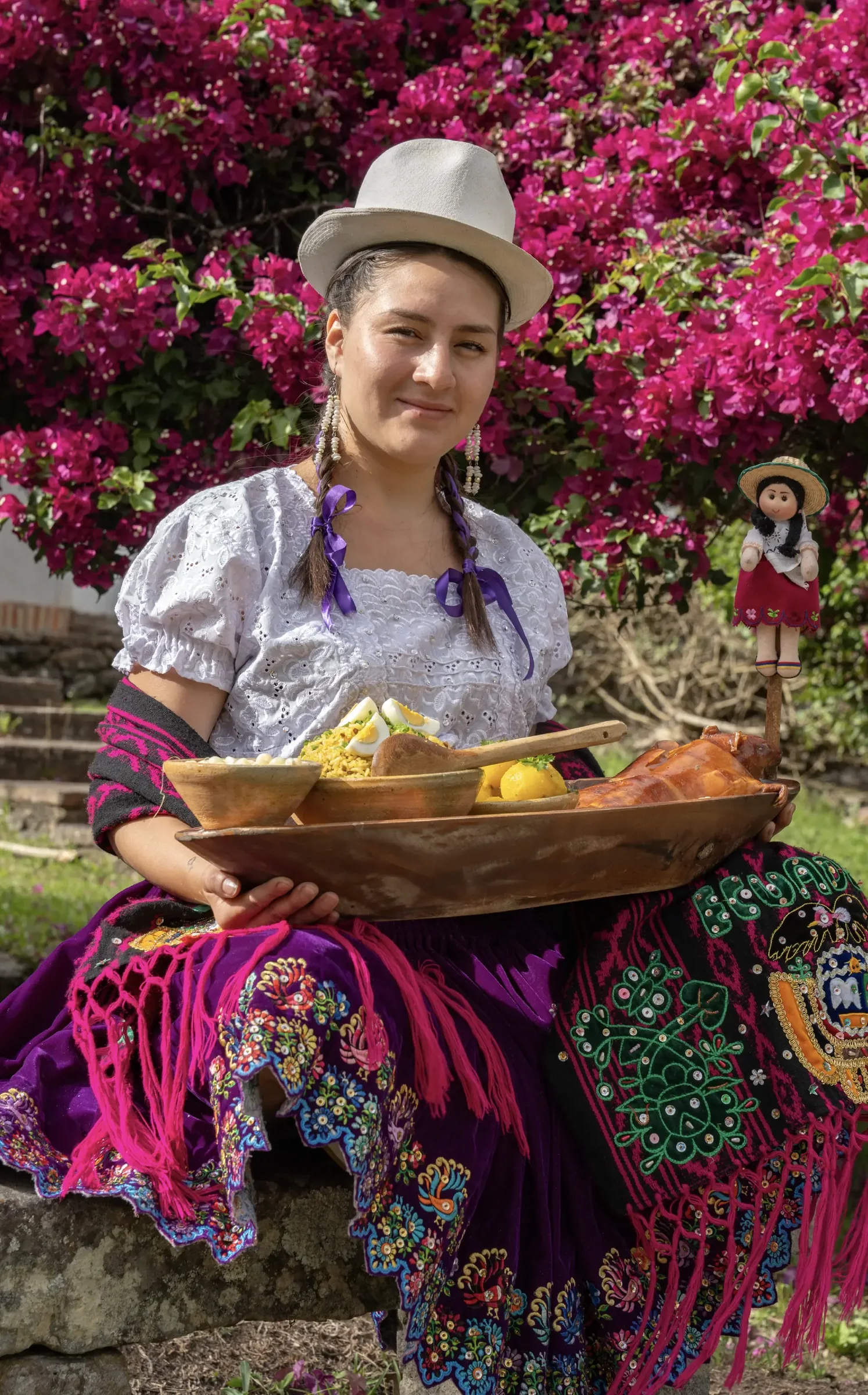 Mujer con vestimenta tradicional sosteniendo una bandeja de comida típica de PauTi, frente a flores rosadas